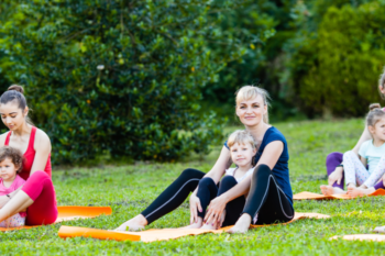 a lady sat on a yoga mat in a field with a child sat between her legs. They are both looking and smiling at the camera