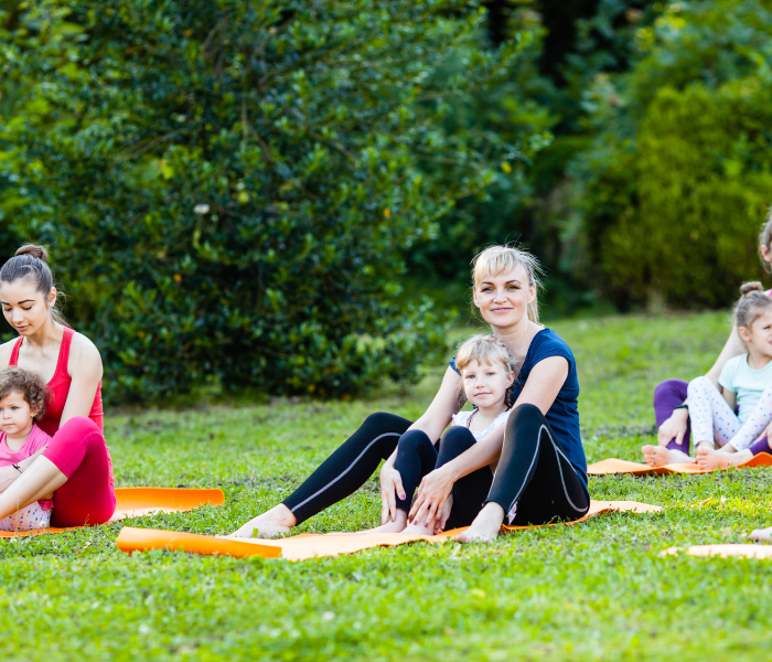 a lady sat on a yoga mat in a field with a child sat between her legs. They are both looking and smiling at the camera