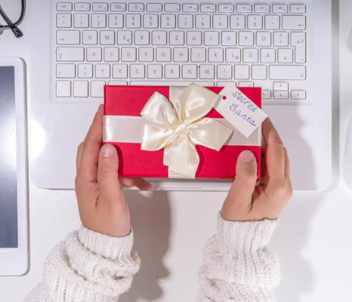 a pair of hands holding a gift wrapped in red paper with a white bow and a label saying secret santa