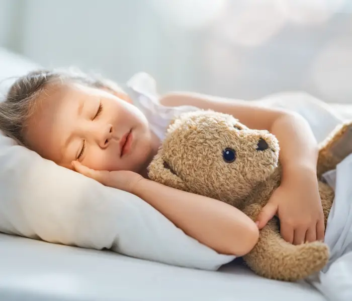 child sleeping on a white pillow, holding a stuffed teddy