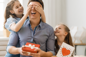 fathers day. a man is holding a gift smiling whilst one girl sits next to him with a card and another girl is covering his eyes