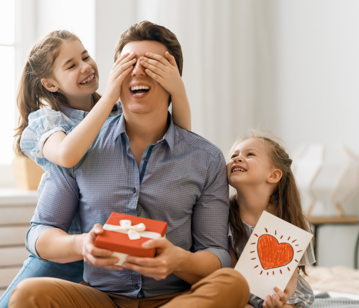 fathers day. a man is holding a gift smiling whilst one girl sits next to him with a card and another girl is covering his eyes