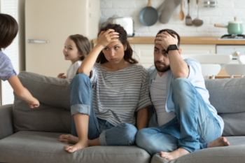 a man and woman sitting on the sofa looking stressed with their heads in hands whilst kids run around behind