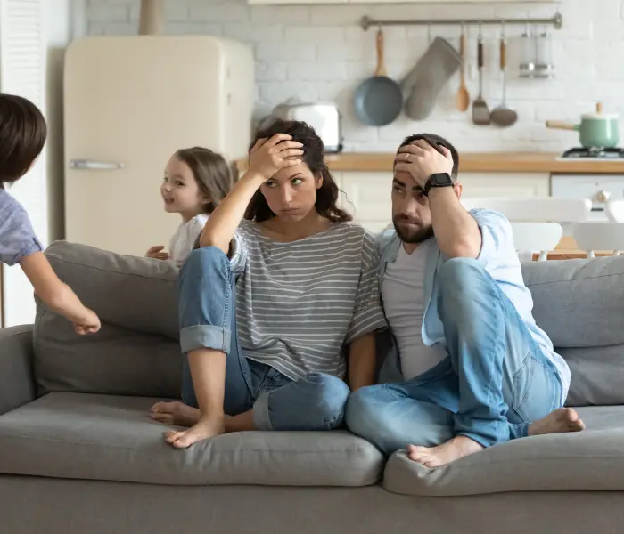 a man and woman sitting on the sofa looking stressed with their heads in hands whilst kids run around behind