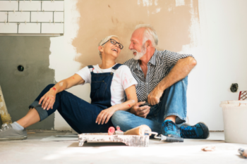 a man and woman sat on the floor with paint in front of them and half painted walls behind them