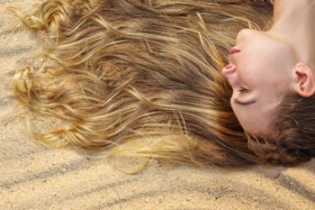 lady with blond hair laid on a beach, you can see the side of her face and her hair is spread along the sand