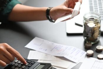 a lady's arms. one hand on a calculator, the other holding reciepts. There are other reciepts on the desk and a jar of coins next to laptop