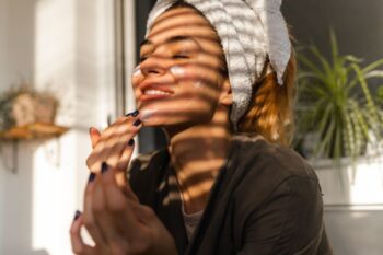 lady smiling in the sunight coming through blinds, she is applying face cream and has a towel over her hair