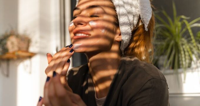 lady smiling in the sunight coming through blinds, she is applying face cream and has a towel over her hair