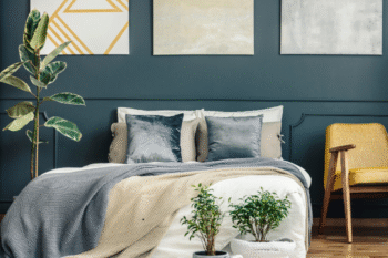 double bed with white bedding and blue cushions and throws. white and gold pictures above and plants in the room