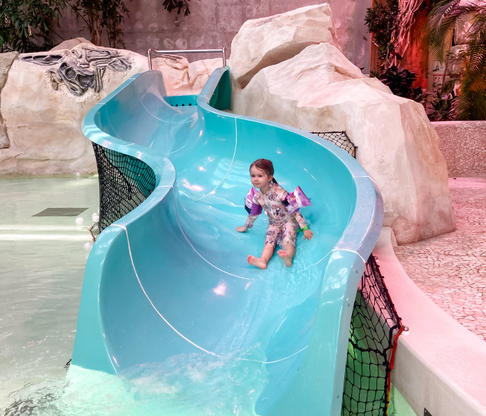 A young girl sliding down a bright blue water slide at Center Parcs Les Landes de Gascogne, surrounded by rocks and tropical plants, during a family weekend.