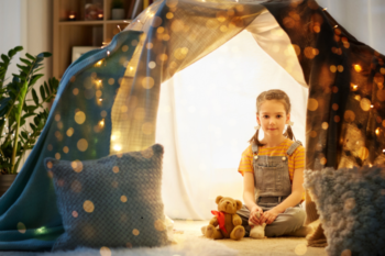Enchanting indoor play tent with a smiling girl and cuddly teddy bear, ideal for kids' imaginative play and family fun. Soft cushions, fairy lights, and a cosy atmosphere create a perfect space for children to relax and enjoy.