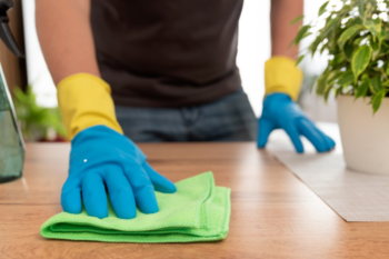 A person wearing yellow and blue gloves cleaning a wooden surface with a green cloth, emphasizing the importance of regular home upkeep.
