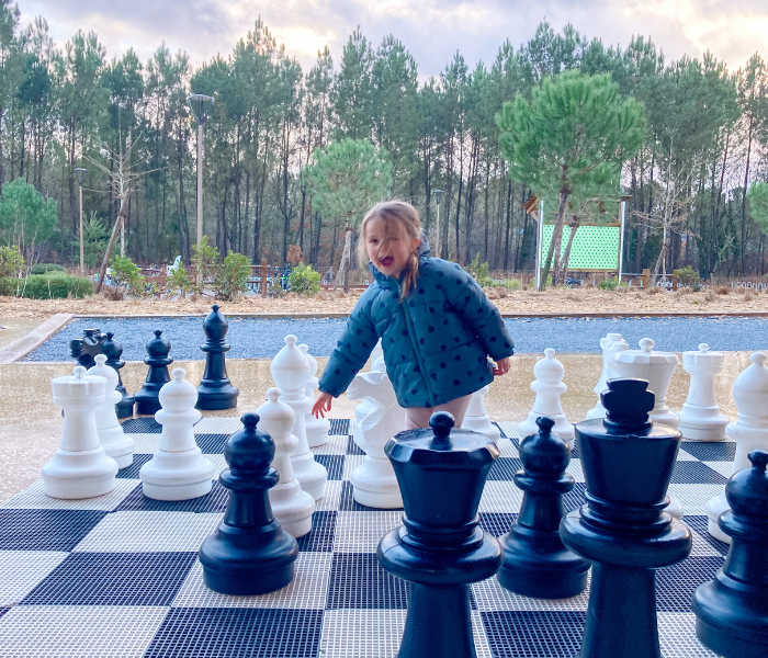 Child enjoying a game of chess on an outdoor giant chessboard at Center Parcs Les Landes de Gascogne, surrounded by nature and trees, during a family weekend.