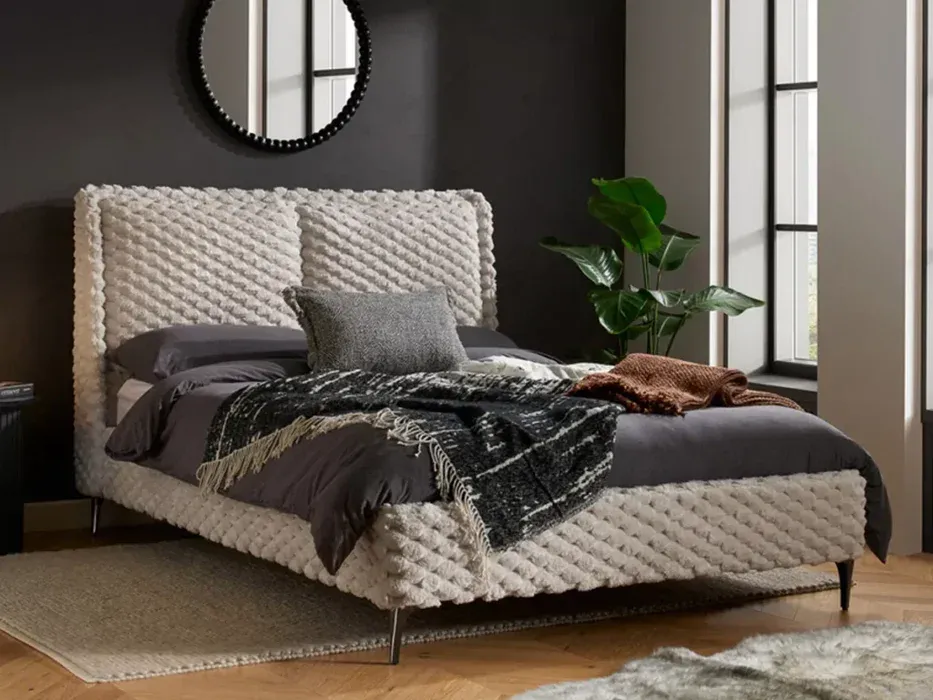 A stylish bedroom featuring a plush, textured bed frame with neutral bedding, complemented by a large green plant and a round mirror, creating a contemporary and inviting space.