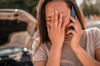 Frustrated woman with long brown hair covering her face, talking on her mobile phone beside a car with an open hood, experiencing a vehicle breakdown on the roadside.