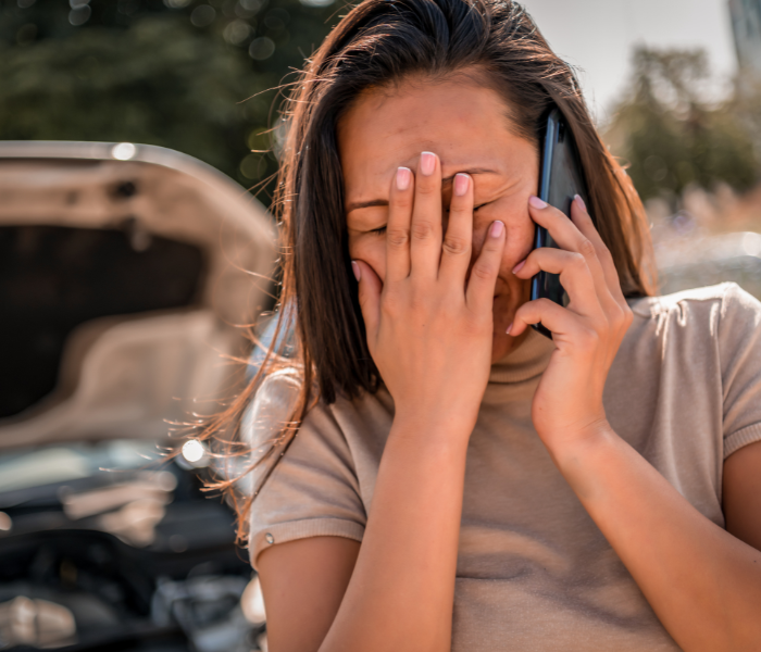 Frustrated woman with long brown hair covering her face, talking on her mobile phone beside a car with an open hood, experiencing a vehicle breakdown on the roadside.