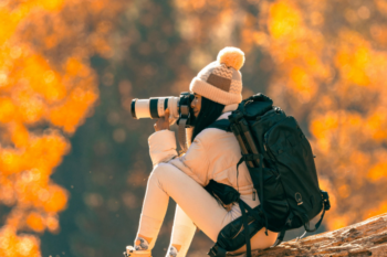 Woman with a camera sitting on a log in autumn forest, capturing nature's beauty with a professional camera, wearing warm clothing and a backpack.