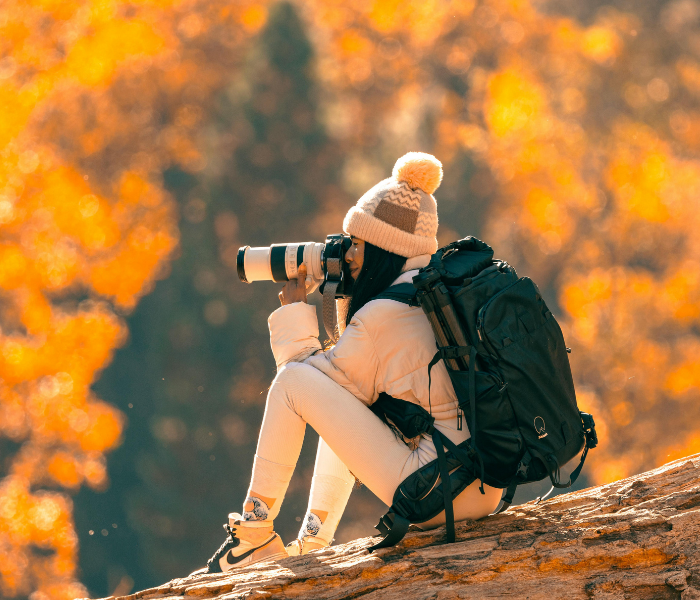 Woman with a camera sitting on a log in autumn forest, capturing nature's beauty with a professional camera, wearing warm clothing and a backpack.
